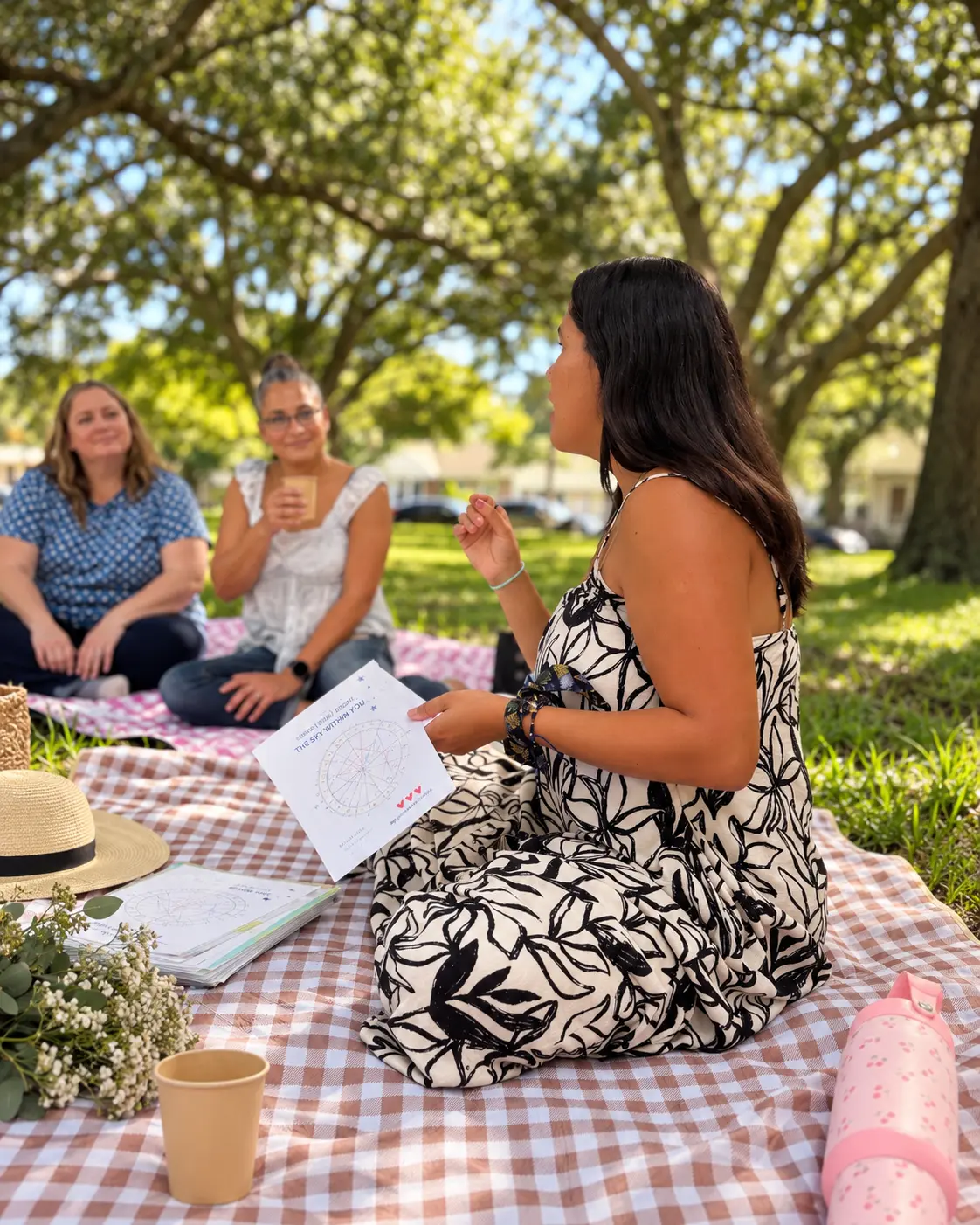 Andrea sitting on a picnic blanket, speaking to a group and holding an astrology chart